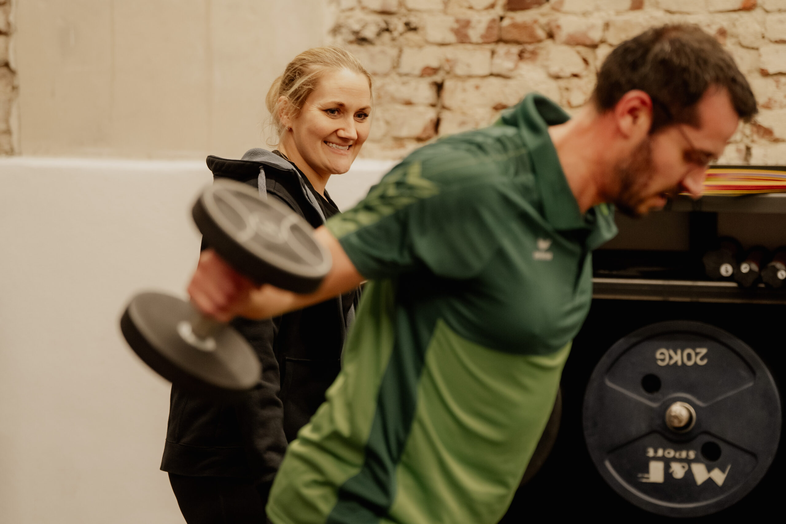 Mann hebt Hanteln beim Personal Teamtraining und Trainerin unterstützt